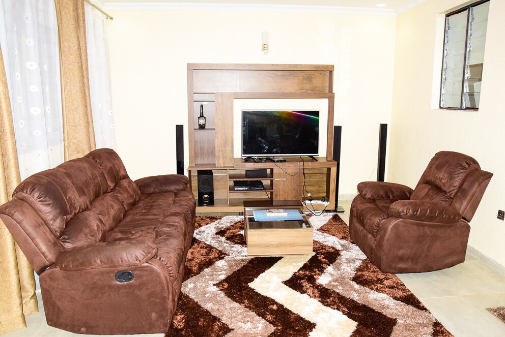 A cozy lounge area features two brown sofas positioned around a coffee table. A television is mounted on a wooden unit against the wall. Light-colored curtains frame the windows, complemented by a patterned rug on the floor.
