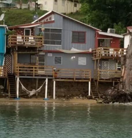 A beachfront studio with two levels is displayed, featuring wooden balconies on both levels. A sandy area is visible at the bottom, with a hammock suspended between the supports. The structure combines blue and red siding, framed by lush greenery in the background.