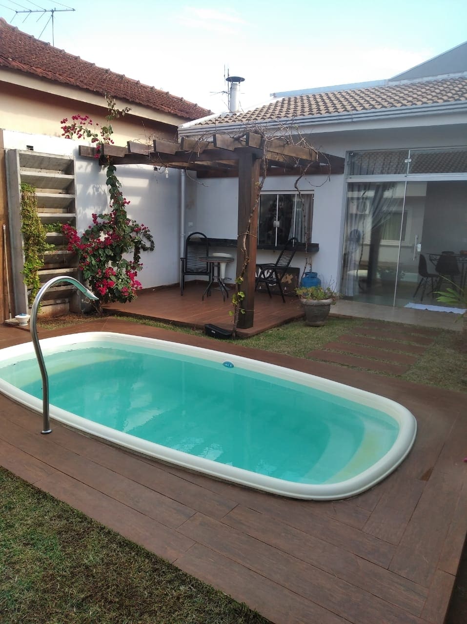 A small swimming pool sits in a well-defined outdoor area, surrounded by wooden decking and patches of green grass. A shaded seating area is visible with a table and chairs, complemented by climbing plants on a trellis nearby.
