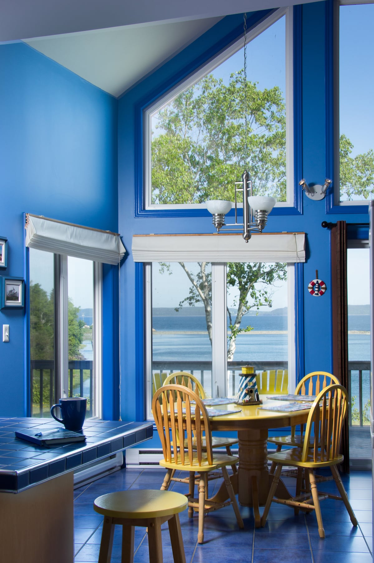 A bright dining area features a round table surrounded by wooden chairs, with large windows offering a view of the ocean and greenery outside. The room is illuminated by natural light, highlighting the blue walls and the modern light fixture overhead.