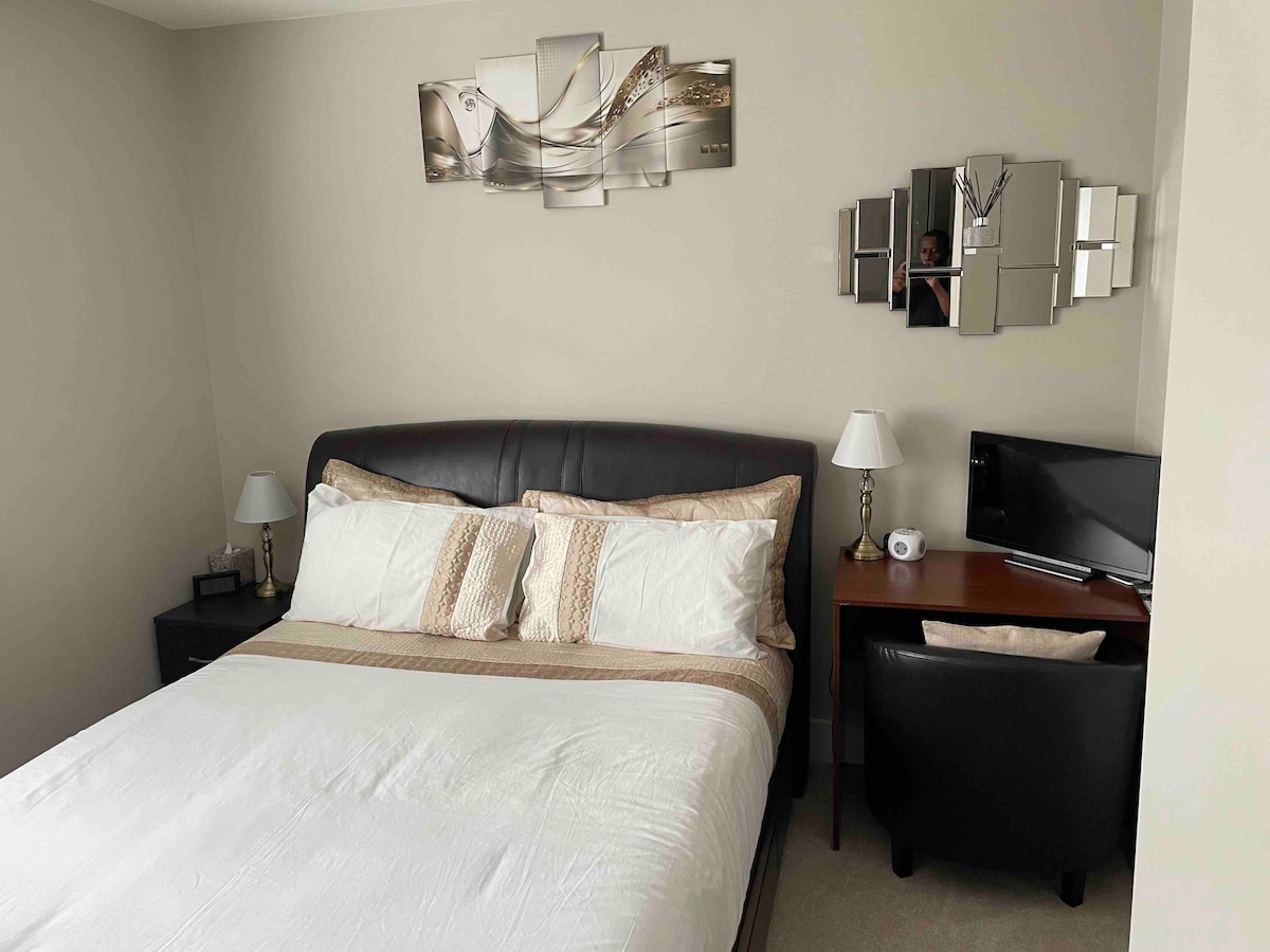 A spacious bedroom features a dark upholstered bed with neutral-toned bedding, complemented by decorative pillows. A mirror wall art piece reflects light. A small desk with a chair and a television are positioned nearby, enhancing the room's functionality.