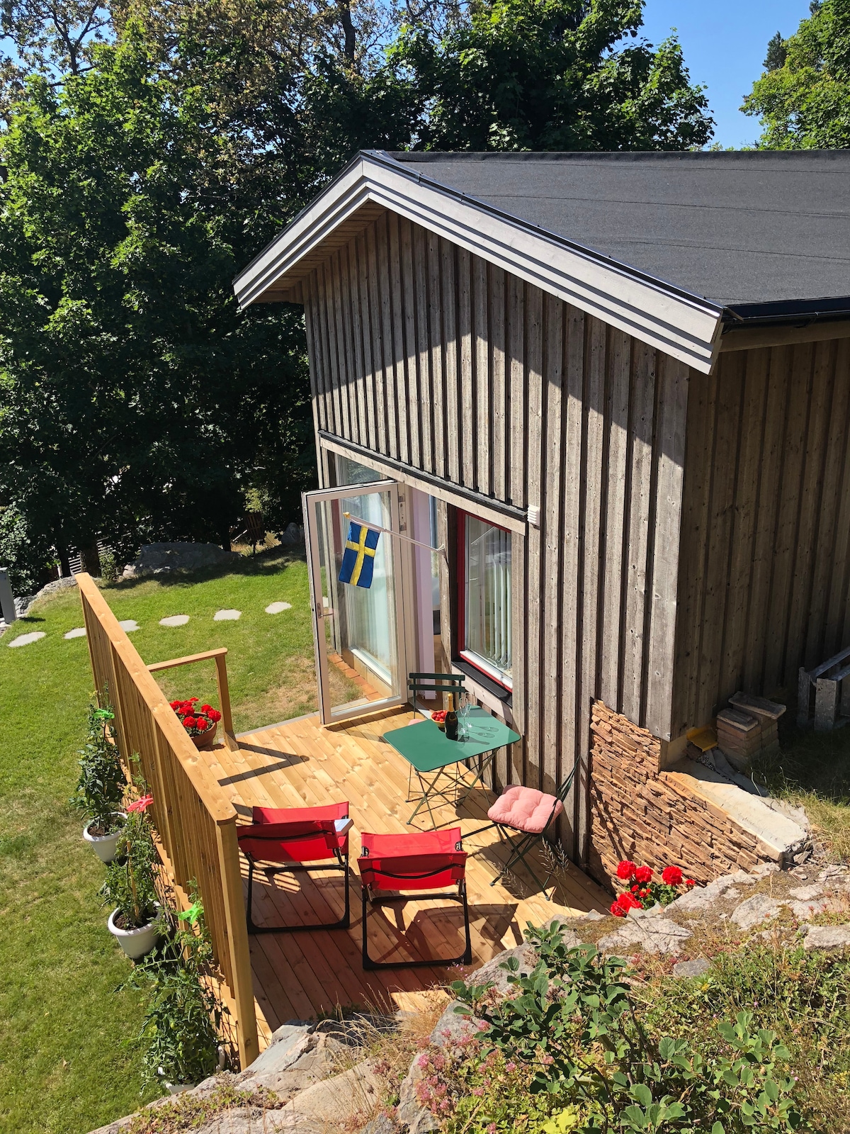An outdoor patio area features a small table and two chairs set against a backdrop of greenery. The building's wooden exterior contrasts with a sunny sky. An open door reveals a glimpse of the interior, and a Swedish flag adds a touch of local charm.