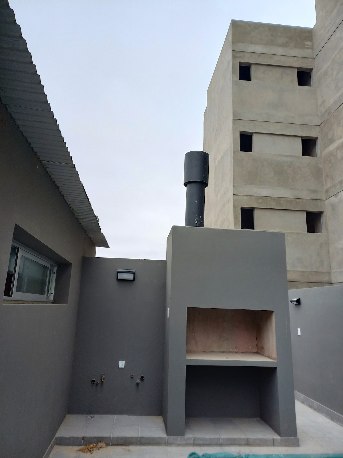 An outdoor area is shown with a built-in barbecue grill set against a gray wall. A tall chimney extends upwards from the grill, while a window is visible to the left. The surrounding area features a paved surface, and a neighboring building can be seen in the background.