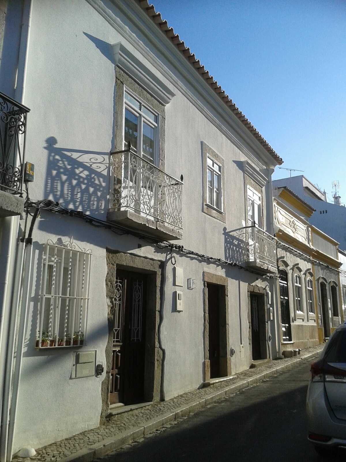 The exterior of a renovated townhouse is displayed, featuring two balconies with ornate railings. The building, painted in light hues, is complemented by traditional stone details and a clear blue sky above. The street is unobtrusive, enhancing the townhouse's charming facade.