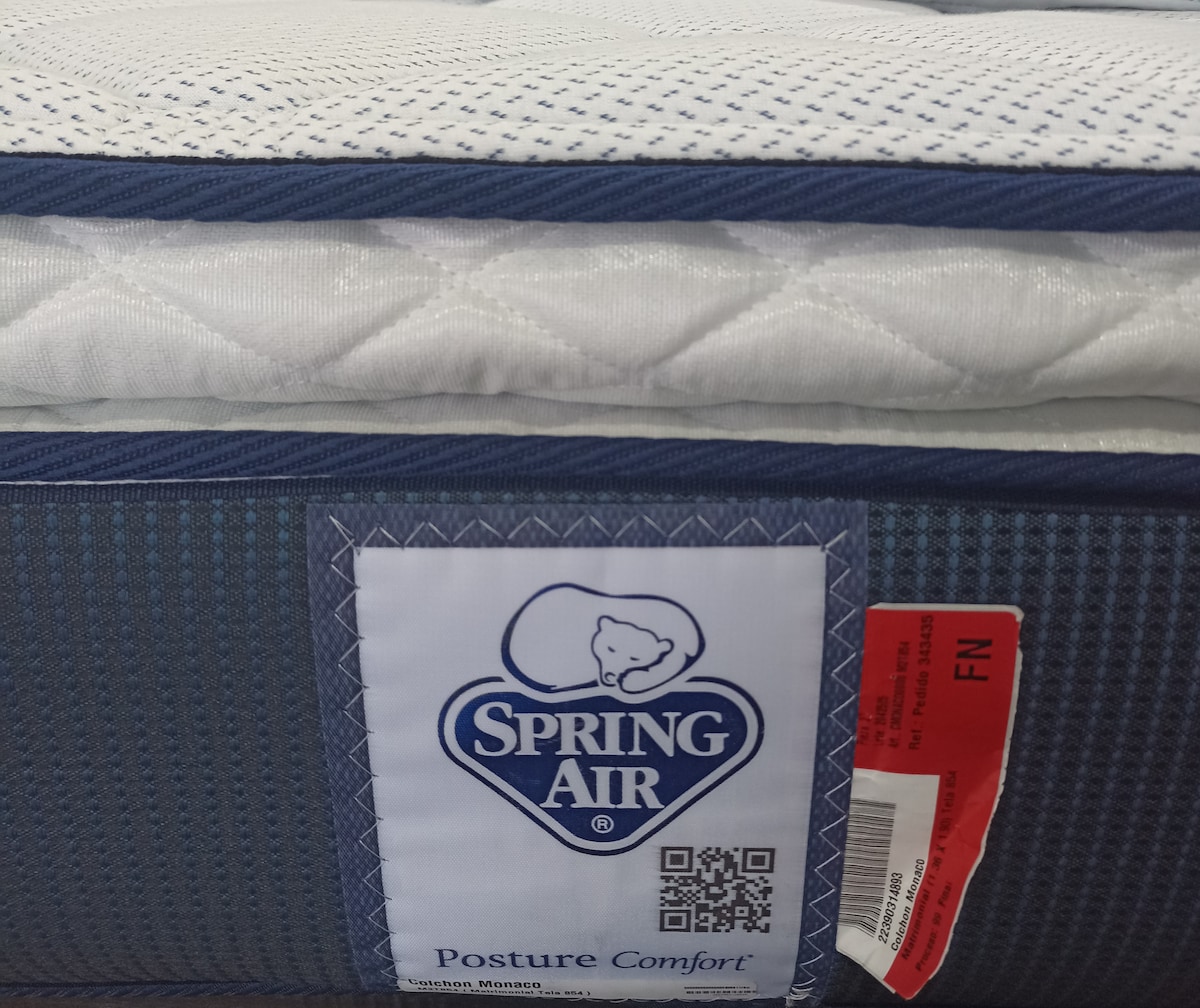 A close-up view of a mattress is displayed, featuring a quilted top surface with a textured blue fabric border. A label is visible, indicating the brand 'Spring Air' along with details about 'Posture Comfort.' The mattress showcases a clean and modern design.