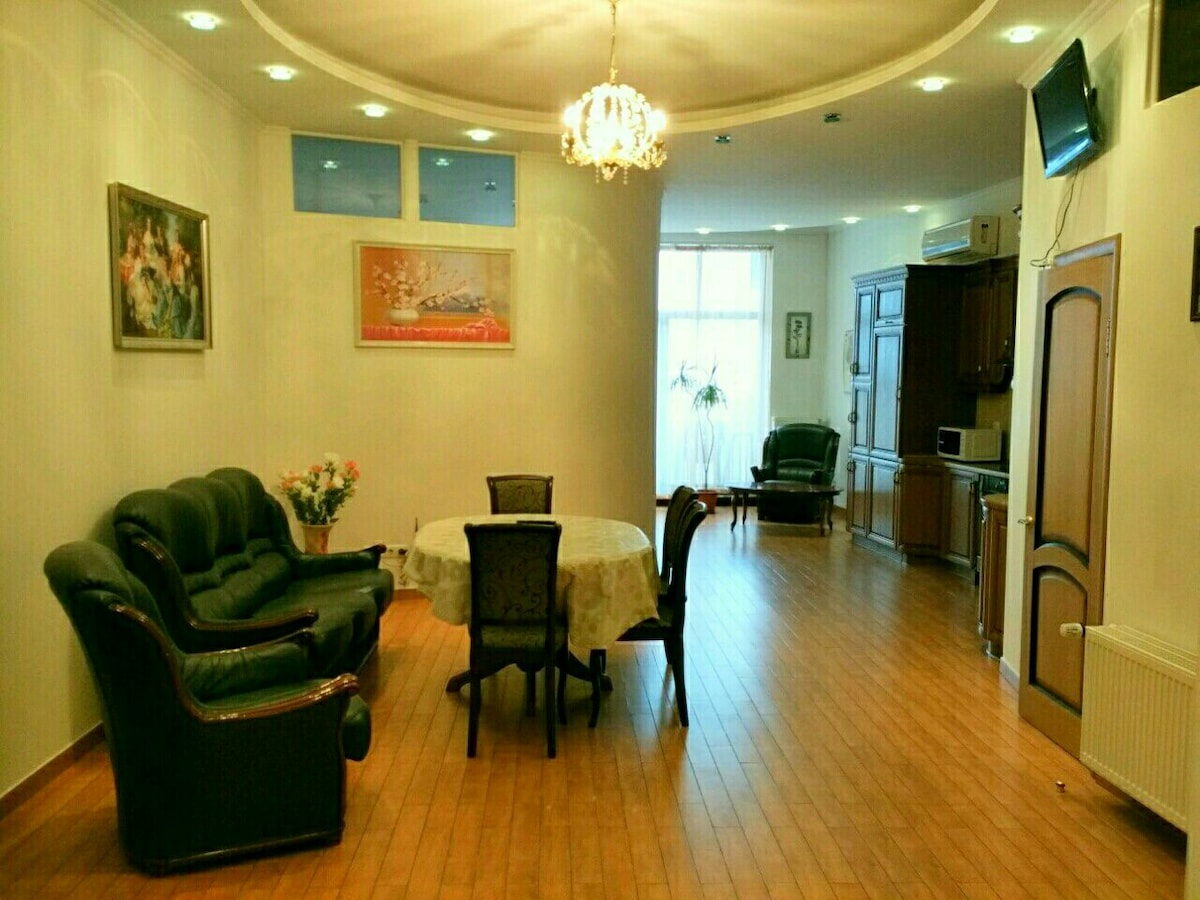 A spacious living area features a decorative chandelier providing soft lighting, surrounded by a green sofa set and a round dining table with six chairs. A kitchenette is visible in the background, and light enters through large windows, enhancing the open feel of the room.