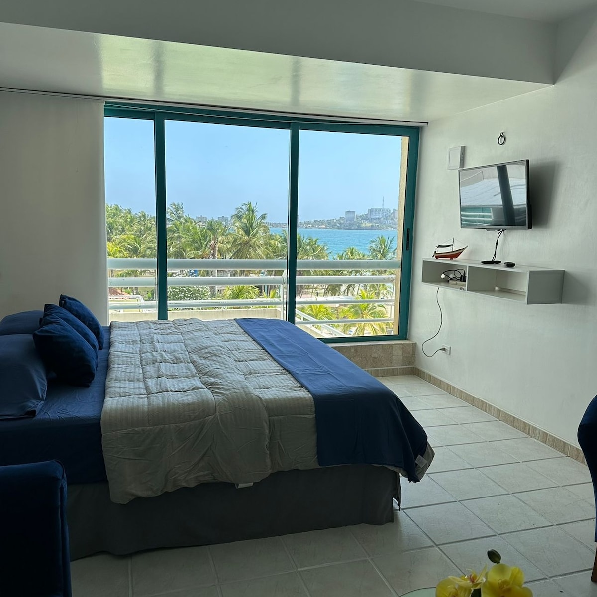 A bright and airy room features a spacious bed dressed in blue linens, positioned next to large glass windows offering a view of the palm trees and ocean beyond. A television is mounted on the wall, and a light-colored floor enhances the room's openness.