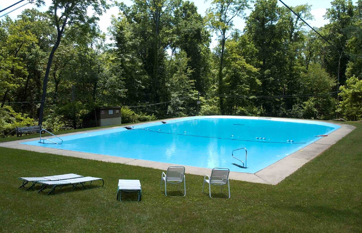 An outdoor swimming pool is surrounded by lush greenery, featuring a bright blue surface. Two white lounge chairs are positioned nearby on a grassy area, while the pool deck has easy access stairs. The tranquil setting offers a serene retreat.