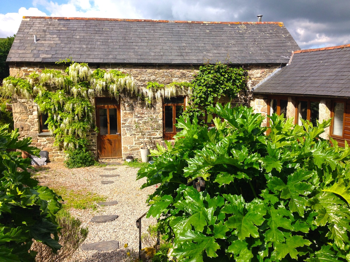 A charming stone building is surrounded by lush green foliage and plants. The exterior features wooden doors framed by climbing vegetation. A gravel pathway leads to the entrance, while tall leaves create a natural border, enhancing the tranquil setting.