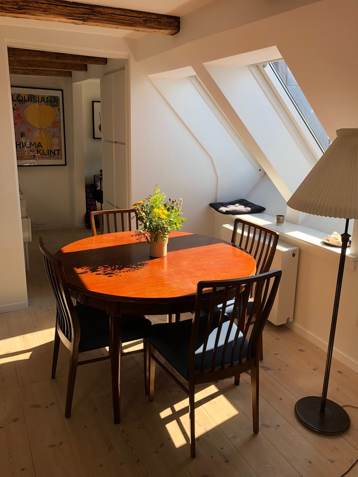 A round wooden dining table is surrounded by four dark wooden chairs, with sunlight streaming through angled skylights. A small vase containing fresh flowers sits at the center of the table, contributing a touch of color to the space.