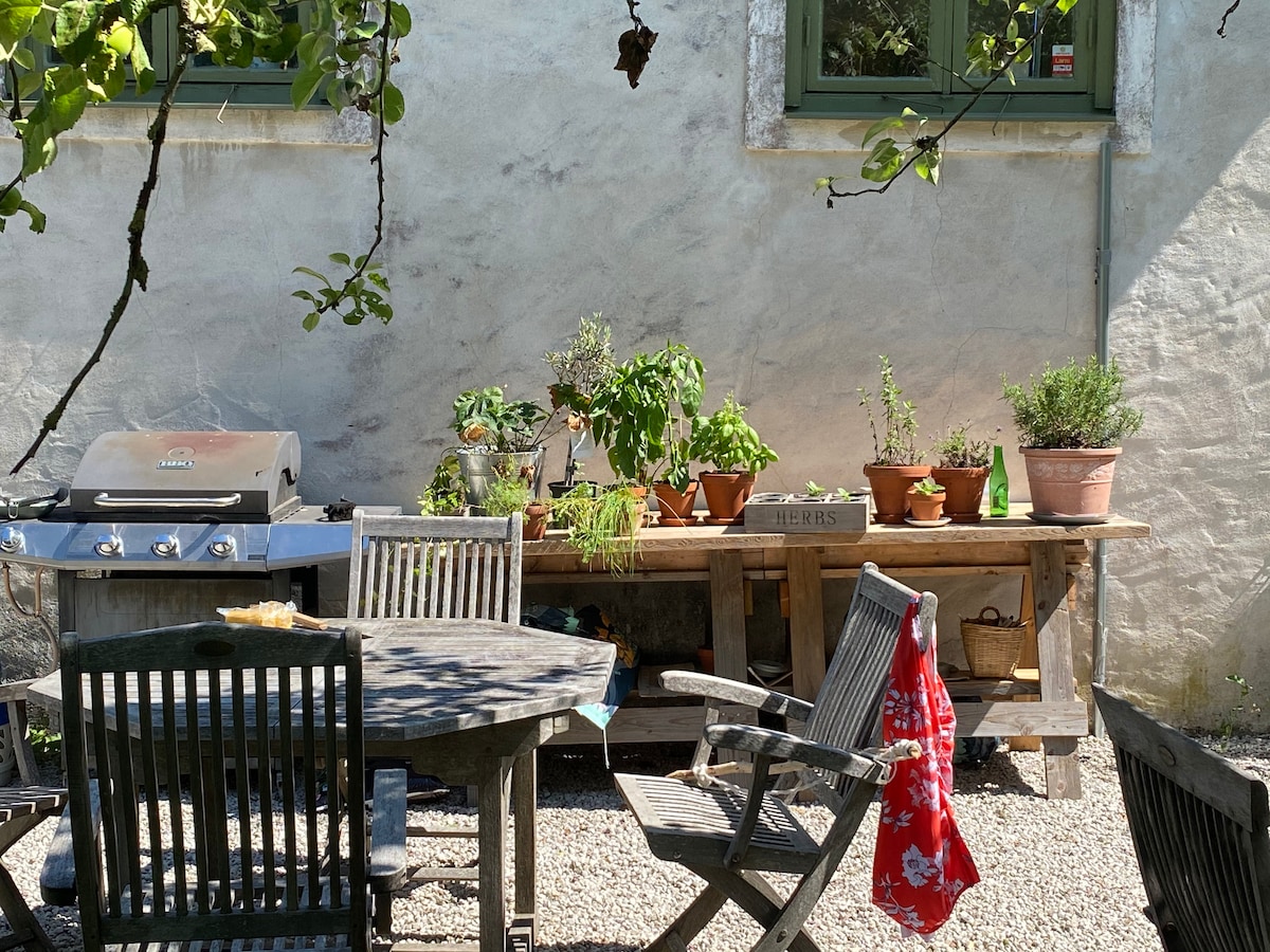 An outdoor space is depicted with a wooden table and several chairs arranged on gravel. A barbecue grill is positioned nearby, and a long table holds potted herbs and plants. A bright red item hangs on one of the chairs, enhancing the setting.