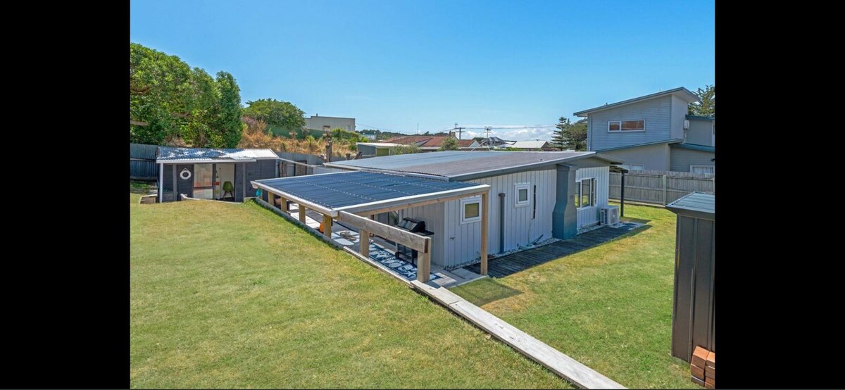 The exterior of the beach house is depicted, showcasing a spacious yard with manicured grass. A covered patio area with seating is visible, alongside solar panels installed on the roof. Surrounding greenery and adjacent structures create a serene atmosphere.