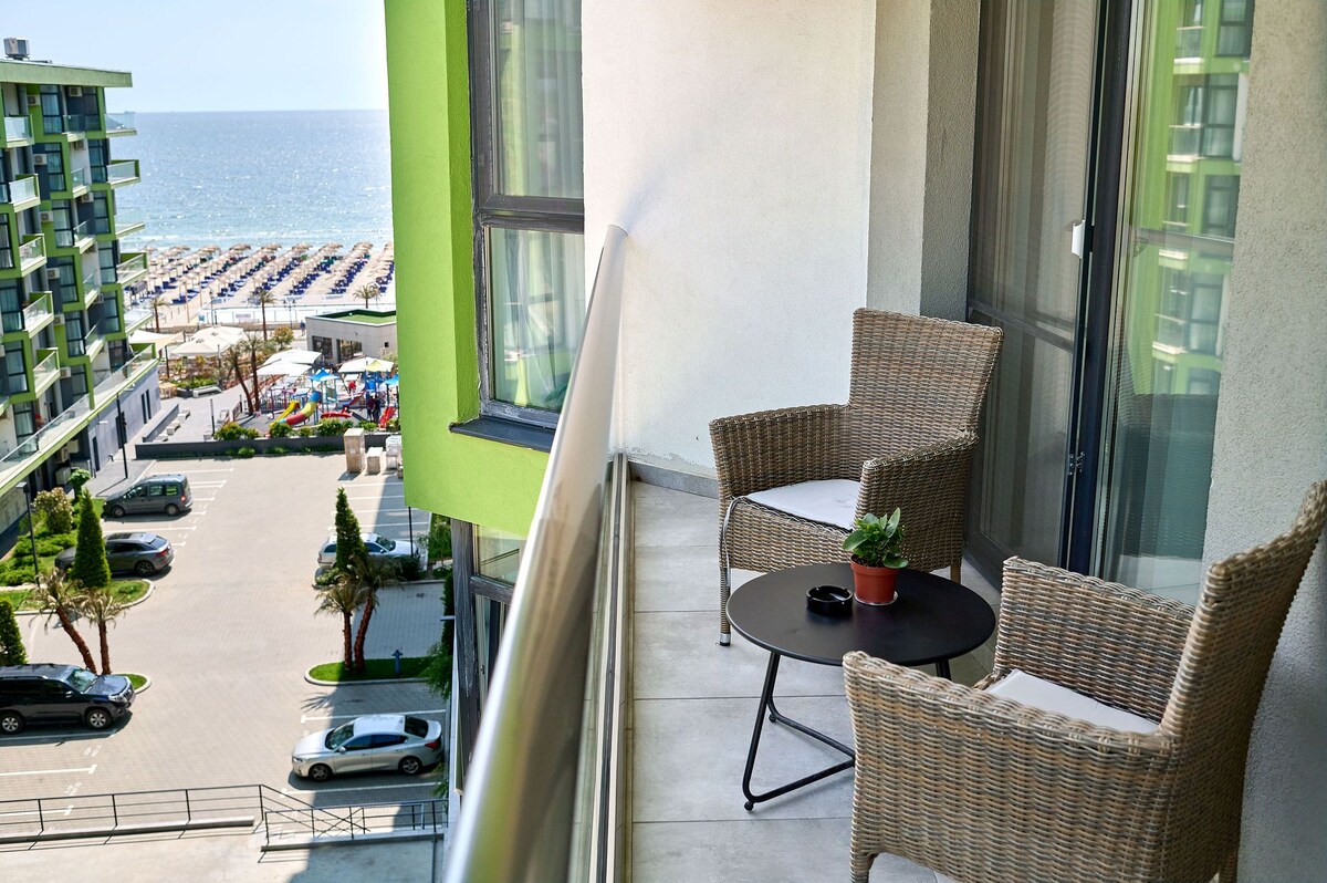 A balcony features two wicker chairs and a small table, accompanied by a potted plant. The view includes sandy beach and ocean, with colorful beach umbrellas and playground visible in the distance. A well-maintained property with parking can be seen below.