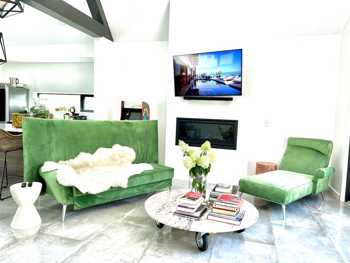 A modern living area features green velvet seating, complemented by a marble coffee table stacked with books. A flat-screen television is mounted on the wall above a sleek fireplace. Natural light streams through the open space, enhancing the relaxed ambiance.