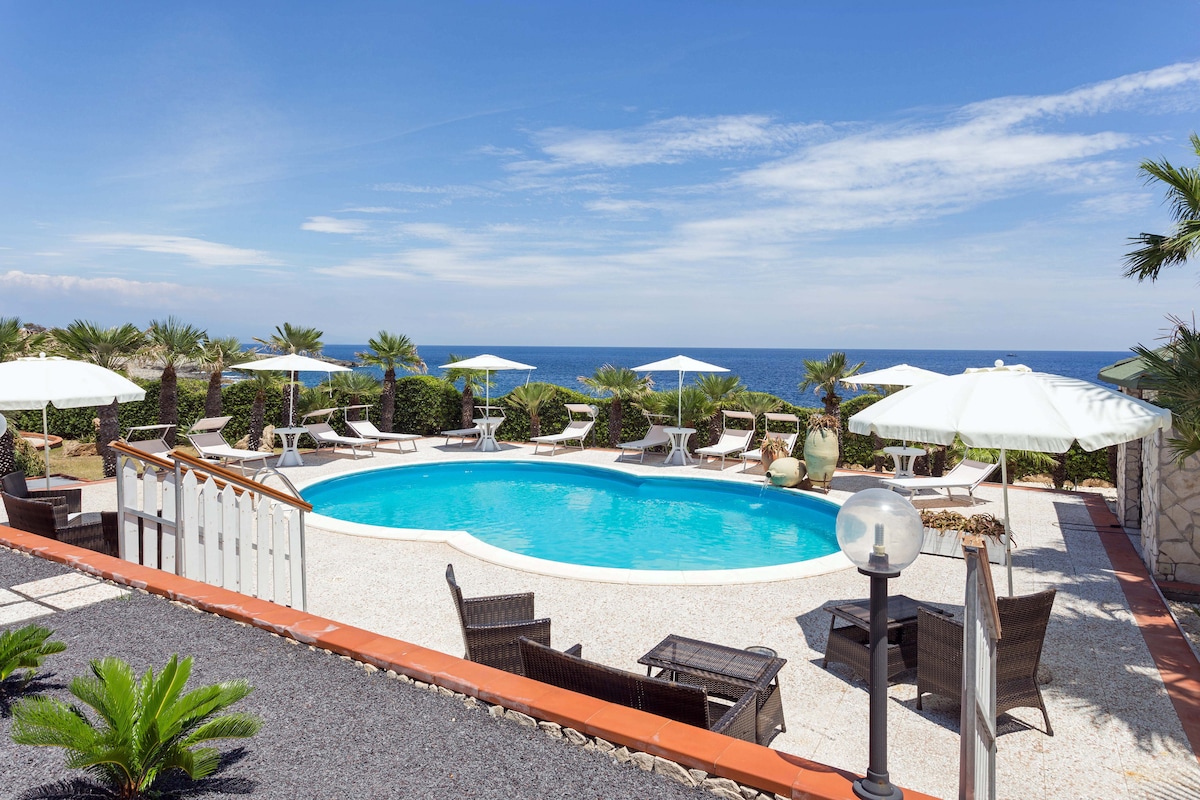 A shared outdoor space features a circular pool surrounded by sun loungers and umbrellas, with views of the sea. Tropical plants are strategically placed around the patio, creating a serene atmosphere. A staircase leads down from a raised terrace to the pool area.