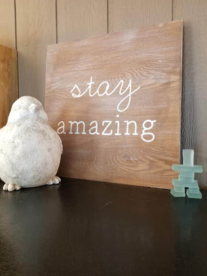 A close-up view of a warm wooden sign displaying the words 'stay amazing' in white lettering. A decorative white bird figurine and a small green sculpture are positioned nearby, adding a charming touch to the cozy interior décor.