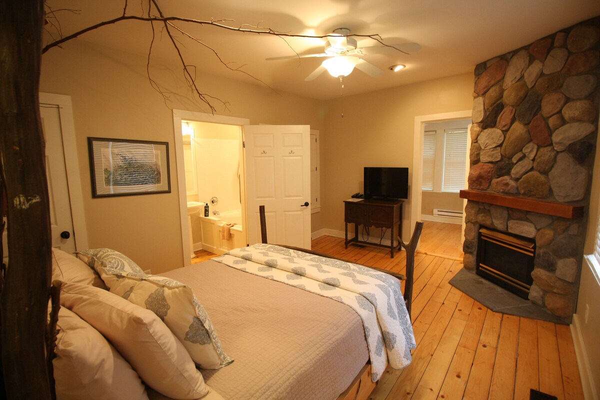 A cozy bedroom features wooden floors and a custom queen-sized bed adorned with a neutral-toned quilt. A stone fireplace adds warmth, while a writing desk and flat-screen TV are positioned nearby. The room connects to an adjoining bathroom, accessible through white-paneled doors.