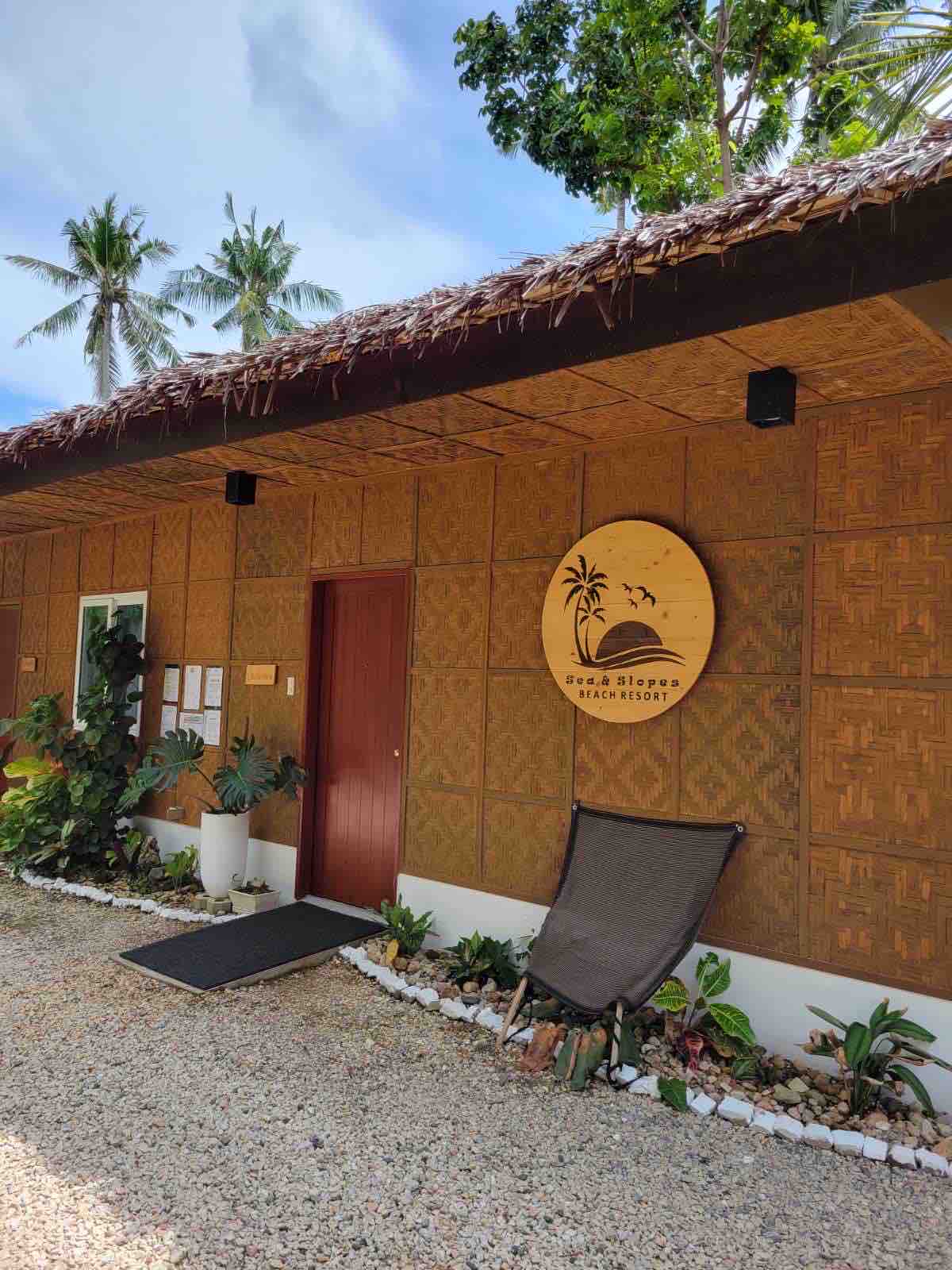 A welcoming entrance to the beach resort is shown, featuring a natural bamboo and wood design. Tropical plants are arranged along the path, leading to a distinct logo on the wall that reflects the coastal environment. A comfortable chair is positioned nearby for relaxation.