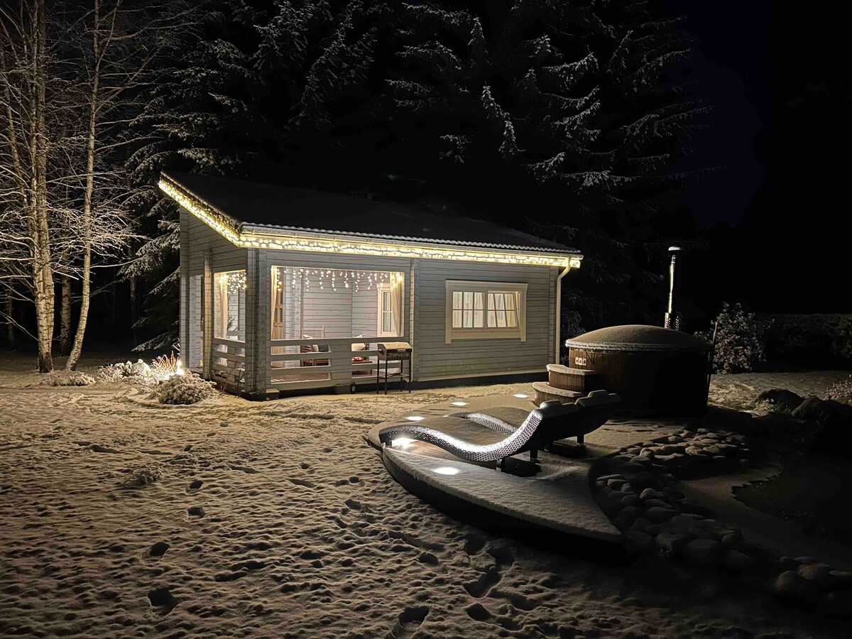 A wooden house is outlined by soft lighting, contrasting with the snowy landscape. A terrace is visible, accompanied by a cozy seating area. The surrounding trees are lightly dusted with snow, enhancing the tranquil winter atmosphere. A hot tub is positioned nearby, partially illuminated.