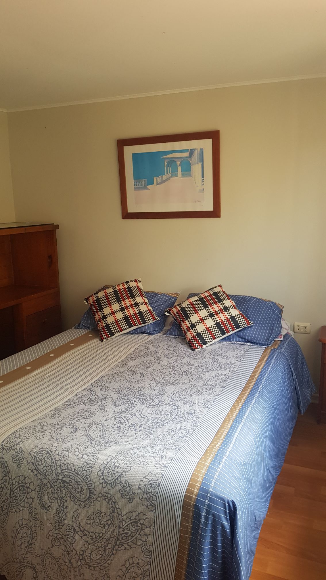 A cozy sleeping area features a spacious bed dressed with a patterned duvet and plaid pillows. A wooden cabinet is positioned nearby, and a framed artwork adorns the wall above the bed, contributing to the room's serene ambiance.