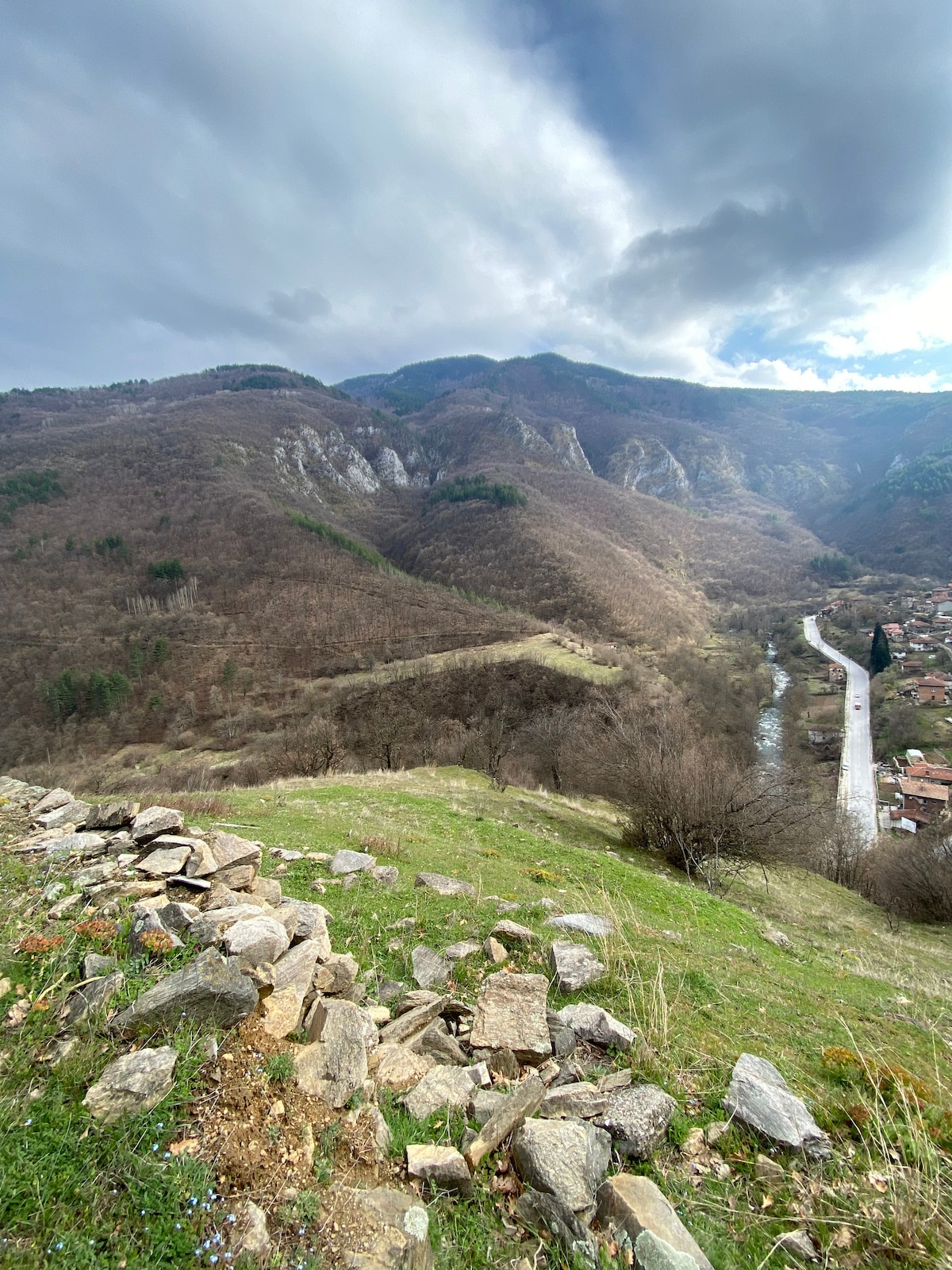 A sweeping view of a mountainous landscape featuring rolling hills covered in brown foliage. A winding river flows through the valley, accompanied by a small village nestled at the foot of the mountains. Cloudy skies create a serene atmosphere over the scene.