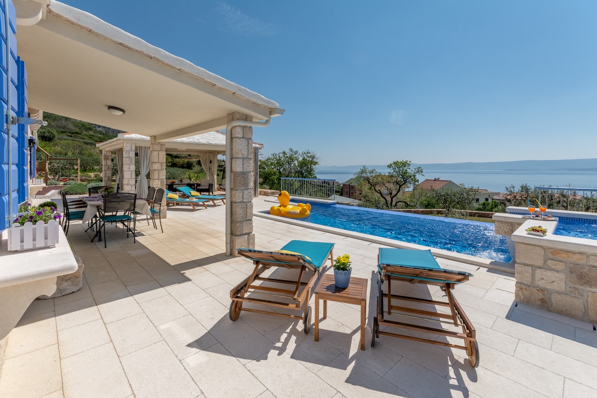 A spacious outdoor terrace features lounge chairs facing a private heated pool. The area is shaded by a pergola, complemented by dining tables nearby. Lush greenery surrounds the space, with clear views of the sea in the background, offering a serene atmosphere.