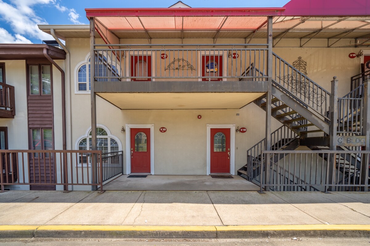 The exterior of the condo features a covered walkway with steps leading to the second level. Two welcoming red doors are visible, accompanied by windows that provide natural light. A railing runs along the walkway, ensuring safety and accessibility.