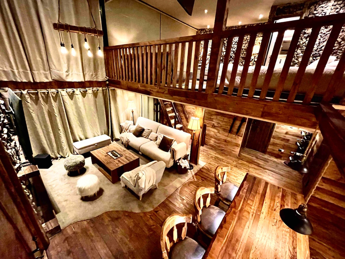 An expansive loft area is presented, featuring a spacious seating arrangement and a wooden coffee table at the center. Soft lighting accentuates the wooden beams overhead, while a cozy dining space with four chairs is positioned nearby. Natural textures and tones create a harmonious setting.