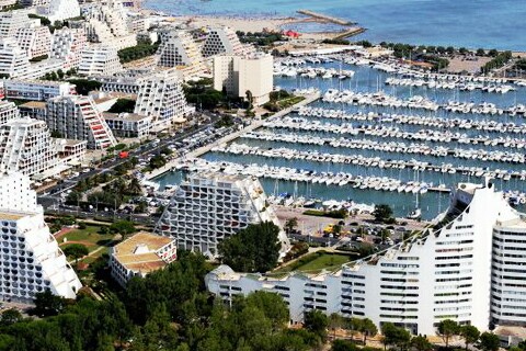 An aerial view captures a marina filled with numerous boats, surrounded by modern architectural structures with distinctive triangular designs. The coastline is visible in the background, flanked by sandy beaches and vibrant waterfront areas, offering a glimpse of a bustling seaside environment.