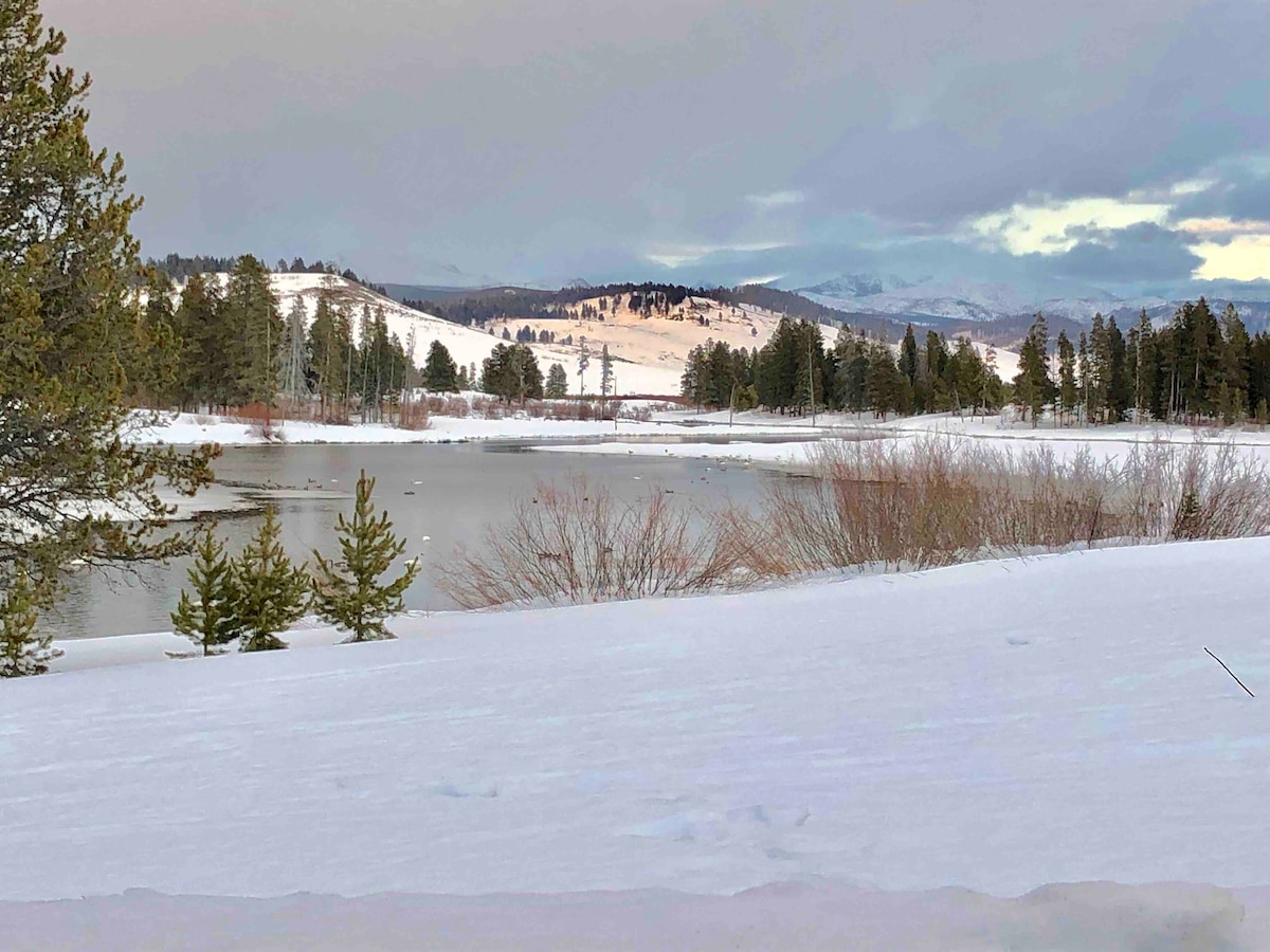 A snow-covered landscape extends towards a tranquil lake, bordered by evergreen trees. In the background, rolling hills and distant mountains are visible under a soft, overcast sky, creating a serene winter scene.