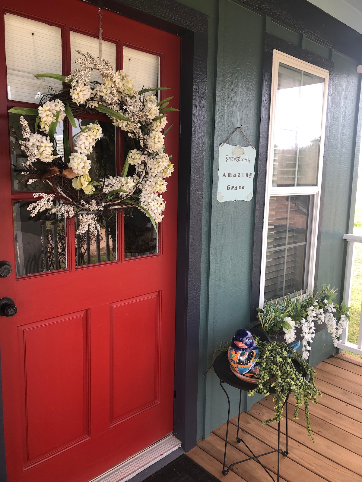 A vibrant red front door is adorned with a floral wreath, evoking a welcoming ambiance. Beside the door, a small table displays a decorative ceramic piece and greenery. A sign labeled 'Amazing Grace' hangs nearby, contributing to the cottage's charming entrance.