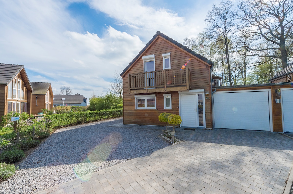 A two-story wooden house is set among greenery, featuring a gravel driveway and a well-maintained garden. The upper balcony provides an outdoor space, while a garage is positioned to the right. Sunlight highlights the house, enhancing the natural surroundings.