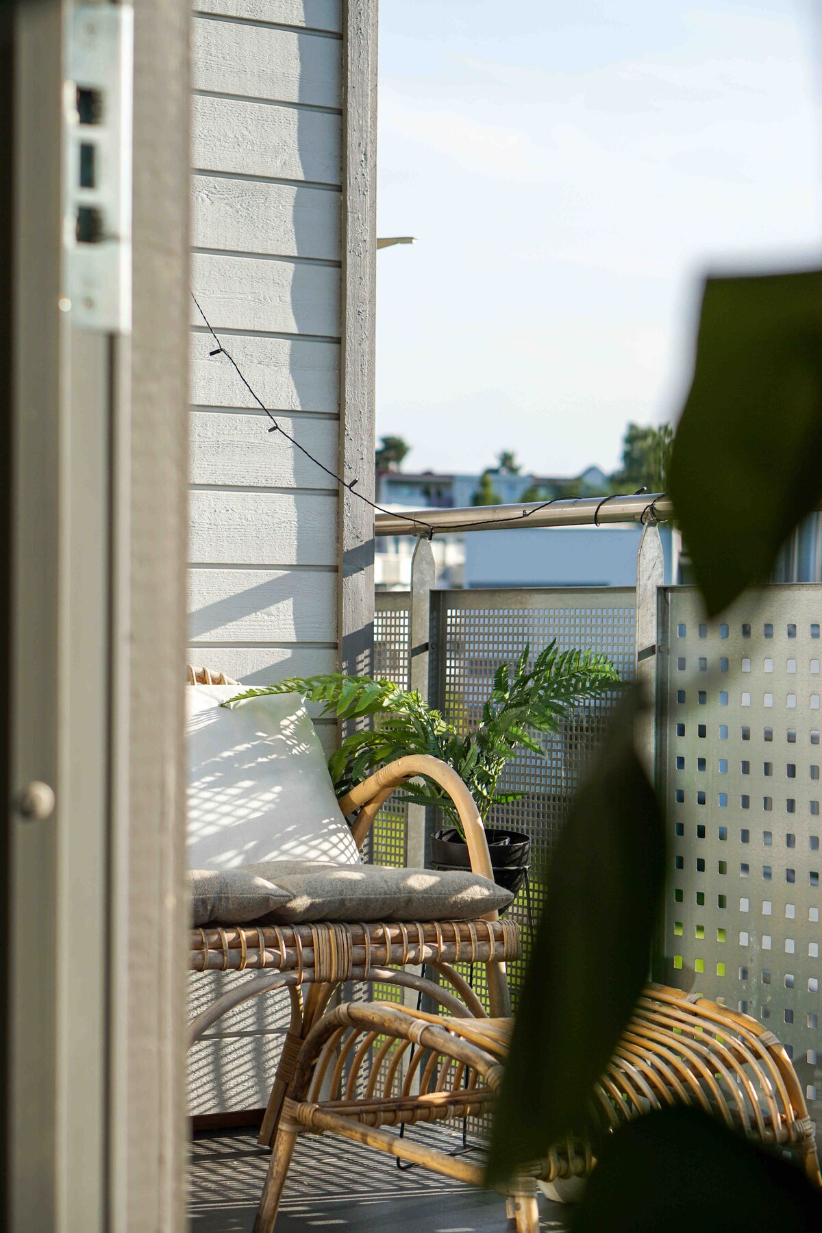 A private balcony is depicted, featuring a wicker chair with a plush cushion, surrounded by greenery. Soft string lights are visible, adding a subtle charm to the space. The view includes distant rooftops under a clear sky.