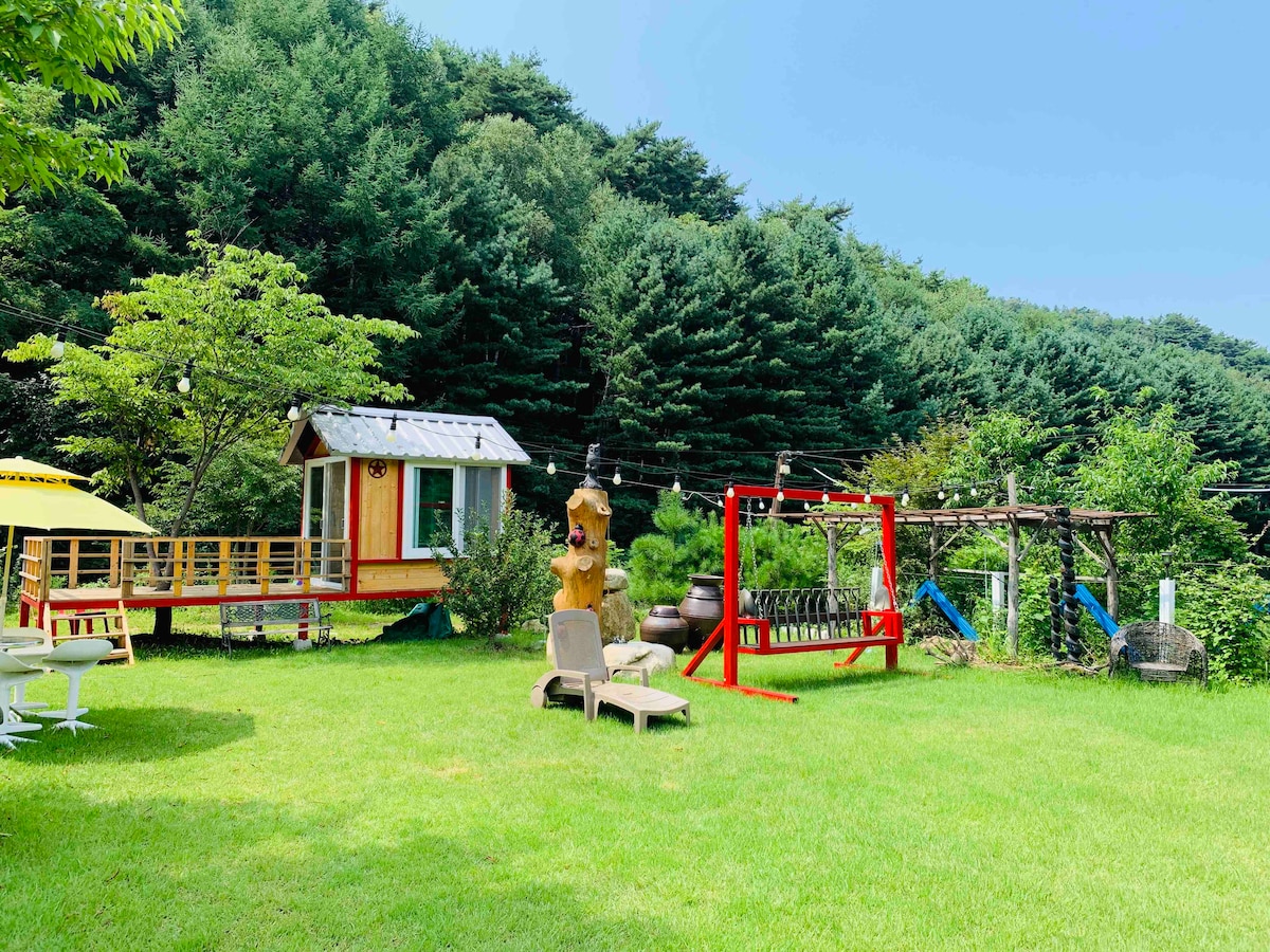 A spacious outdoor garden features green grass and various play structures, including a vibrant red swing set and a wooden playhouse. Surrounding trees provide a natural backdrop, while lounge chairs encourage relaxation in the sun. String lights create a welcoming atmosphere for evening gatherings.