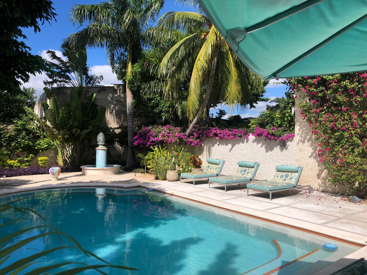 A serene pool area is surrounded by vibrant tropical plants and flowering bougainvillea. Three lounge chairs with soft cushions are positioned by the clear water, while a stone water feature adds a peaceful element to the lush environment.