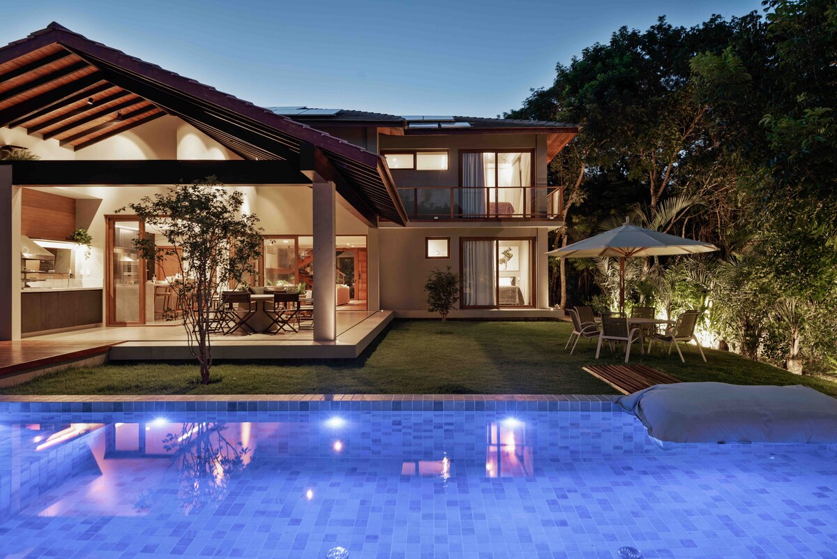 A spacious outdoor area is observed, featuring a well-lit swimming pool with blue-tinted water. The house is designed with large glass doors that open to the patio, where a table and chairs are set up underneath a shaded umbrella. Lush greenery surrounds the scene.