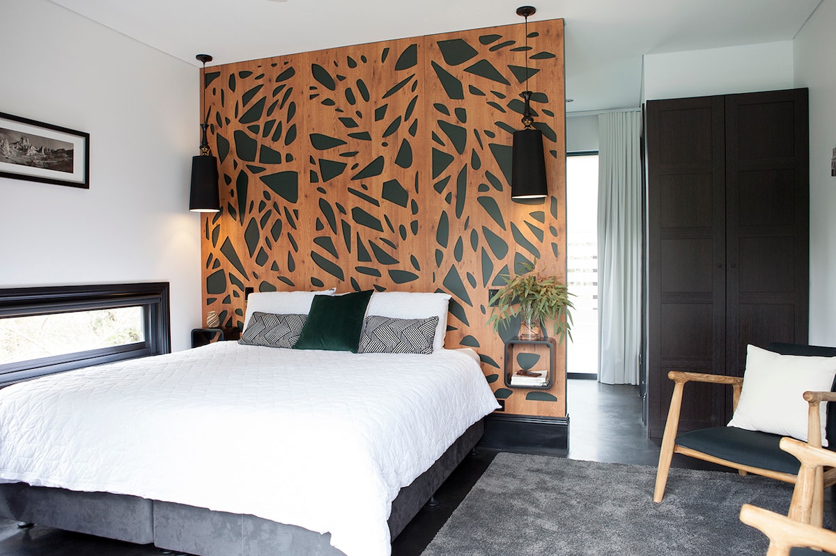 A stylish ensuite guest room features a king-sized bed with a white quilt and a selection of soft pillows. A striking patterned feature wall adds visual interest, while a cozy chair is positioned nearby. Natural light illuminates the space, creating a serene atmosphere.