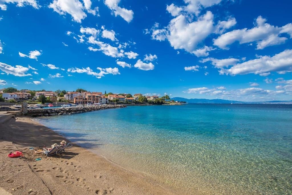 A serene beach scene presents clear blue waters gently lapping against a sandy shore. Distinct buildings line the beach in the background, surrounded by lush greenery, while scattered clouds drift across the bright sky. The tranquil setting invites relaxation and offers scenic views of the distant mountains.