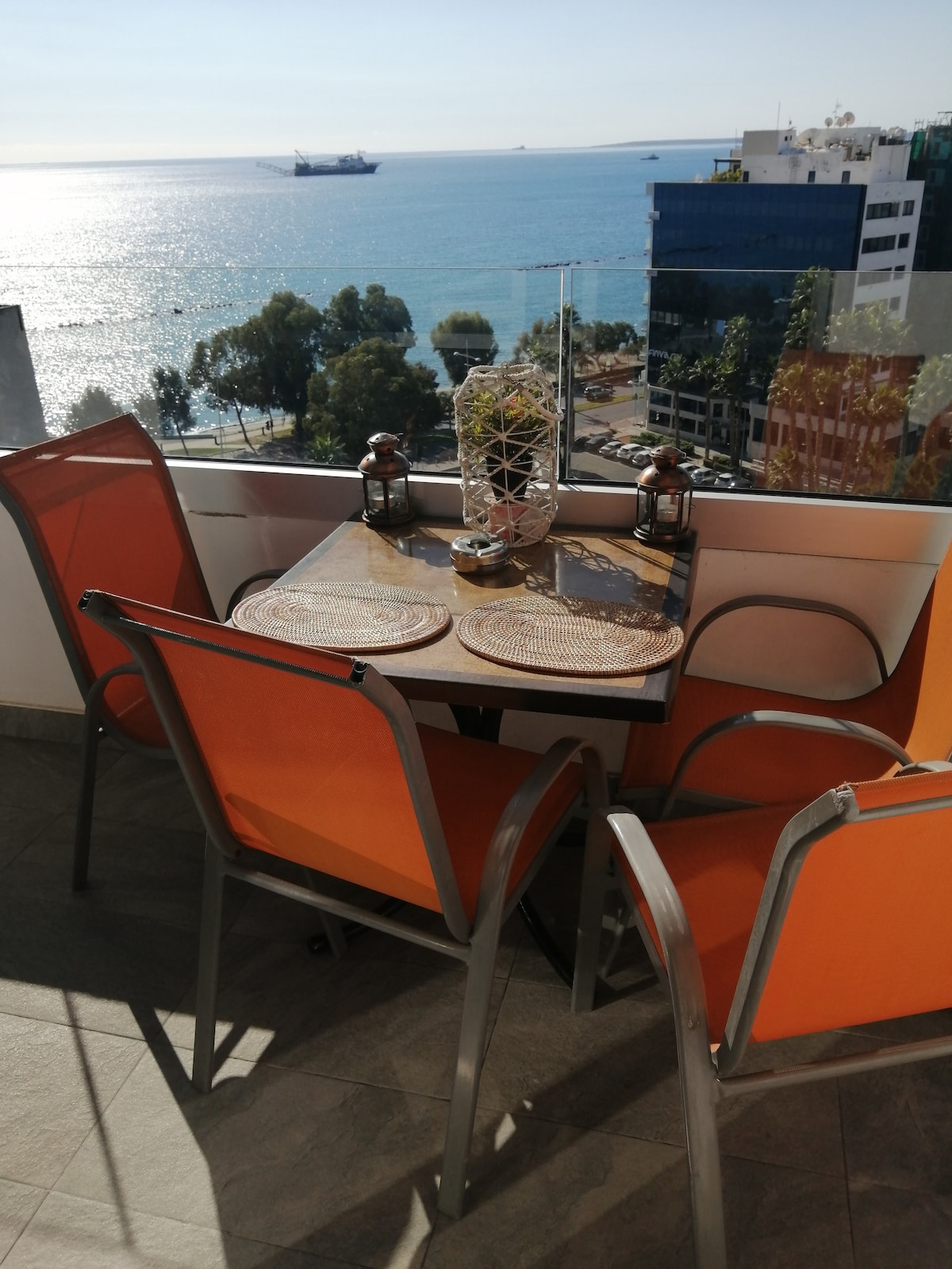 A dining table with two orange chairs is positioned on a balcony, offering views of the sea. Natural light reflects off the water's surface, while a decorative centerpiece and lanterns adorn the table. The surrounding landscape features greenery and urban buildings.