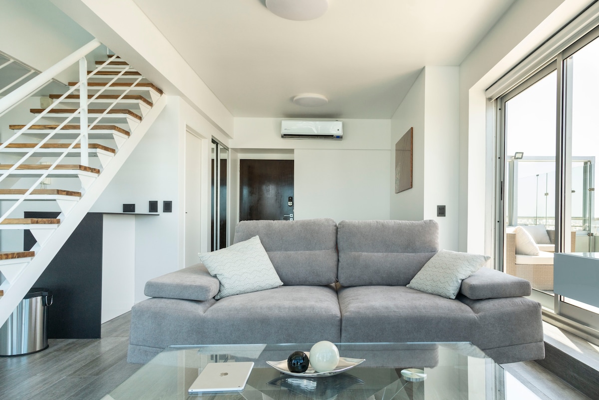 A spacious living area features a comfortable gray sofa adorned with neutral cushions, paired with a glass coffee table. Large sliding glass doors provide abundant natural light and showcase the view of the exterior terrace. A staircase leads to the upper level.