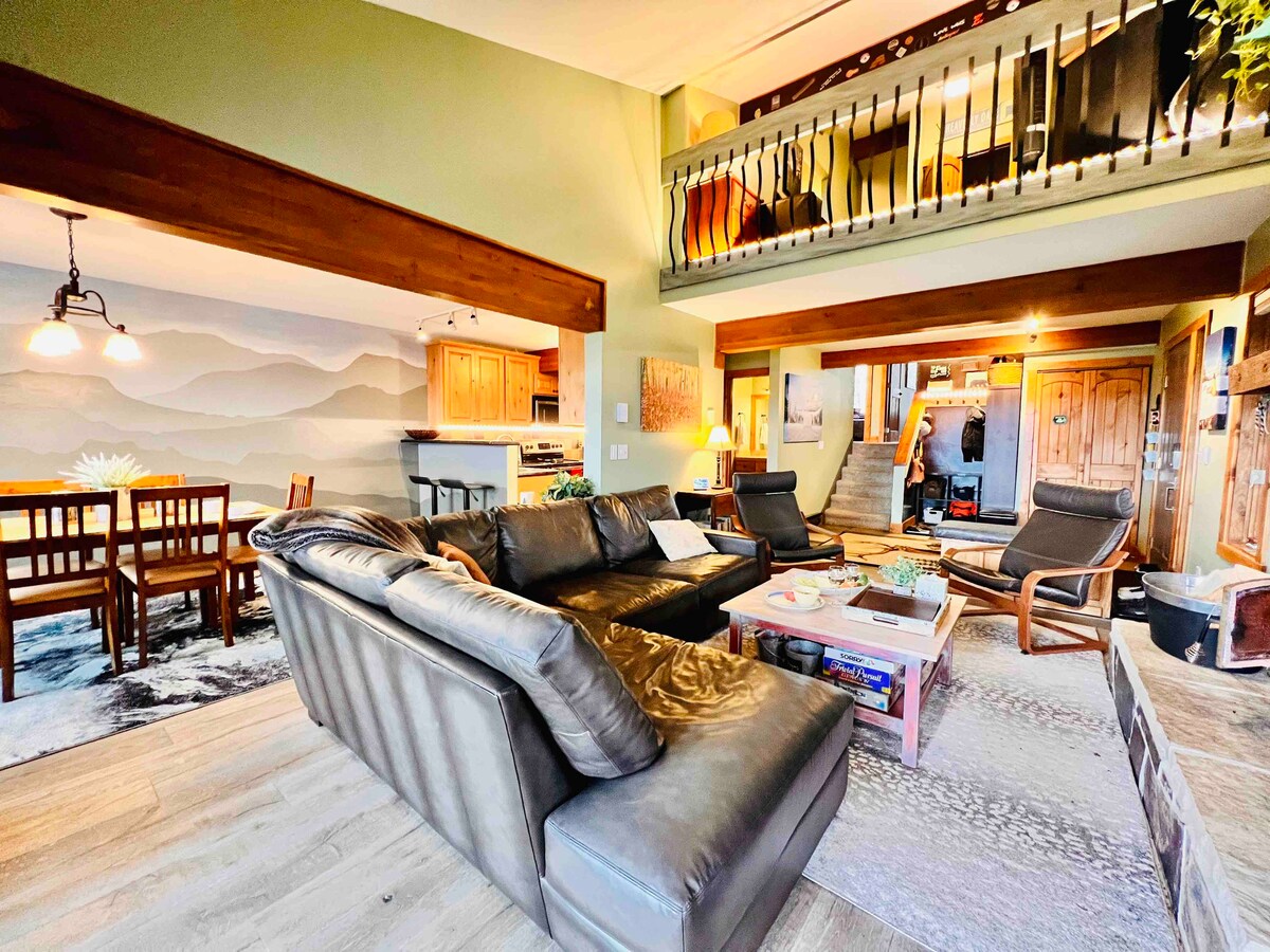 An open living area features a spacious leather sectional sofa facing a coffee table. Natural light fills the space from large windows, highlighting the earthy tones of the decor. A dining area with wooden chairs is visible, along with glimpses of the kitchen and staircase to the upper level.