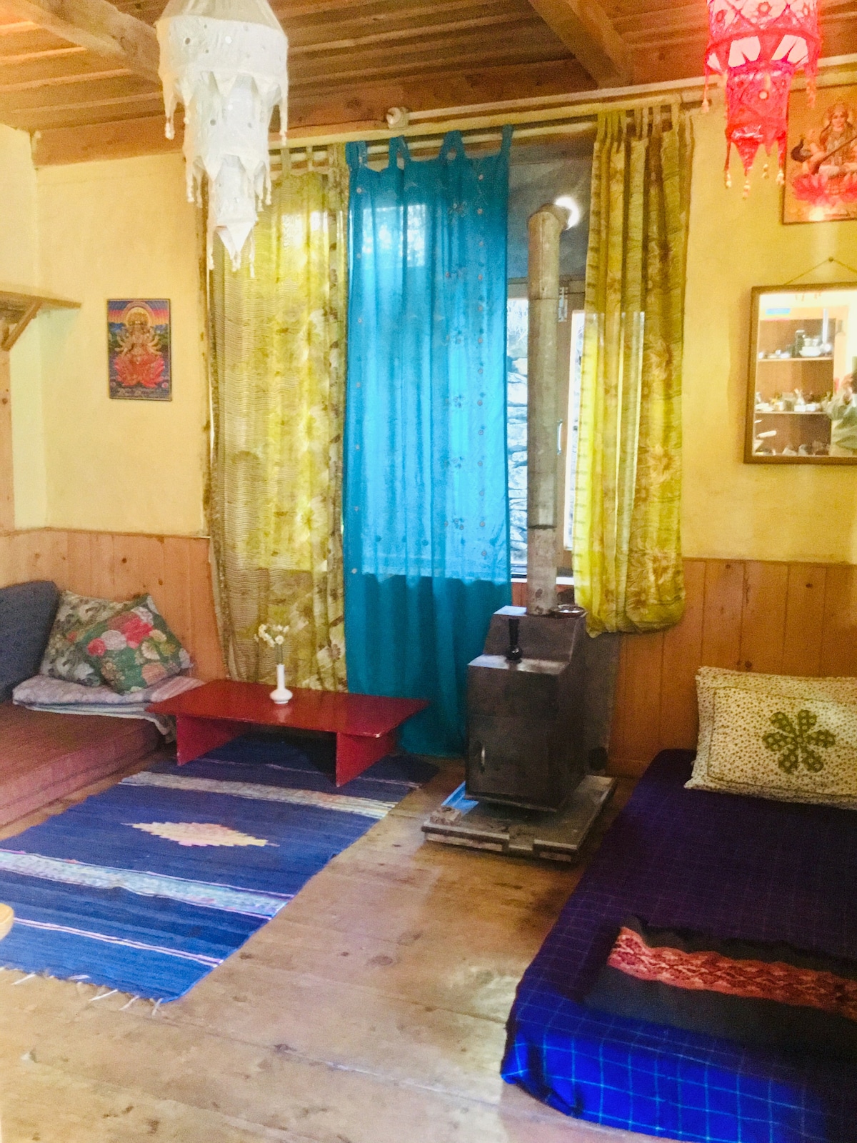 The living area features a warm design with wooden walls and a wooden floor. Colorful curtains frame the window, while pillows are arranged on two low seating areas. A wood-burning stove stands as a focal point, accompanied by a small table and vibrant rugs.