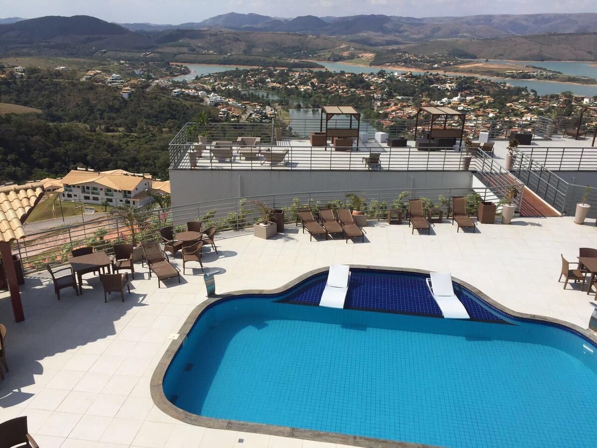 A rooftop area features a refreshing pool with blue tiles, surrounded by lounge chairs. Scenic views of the landscape and the lake are visible in the background, with gentle hills adding depth to the horizon. Greenery is present along the perimeter.