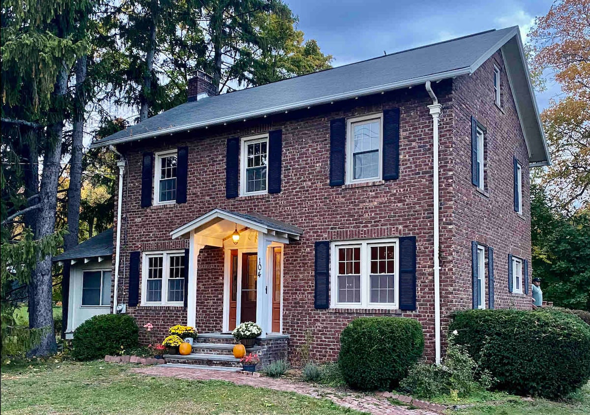 The exterior of a classic brick house is presented, featuring a welcoming porch under a gabled roof. Dark shutters frame the windows, and neatly trimmed shrubs and seasonal decorations enhance the front yard. Tall trees provide a serene backdrop to the property.