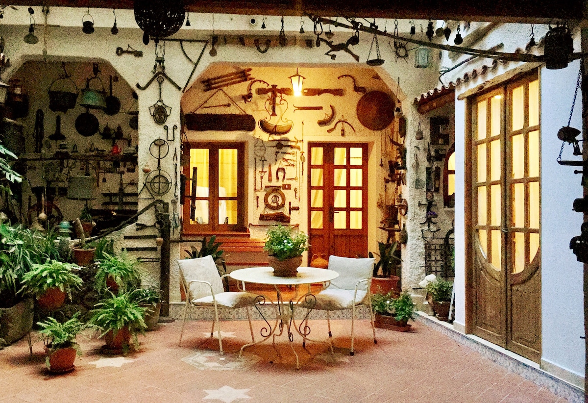 An outdoor courtyard is surrounded by rustic decor and numerous plants, creating a peaceful environment. A round table with two chairs is positioned at the center, while wooden double doors stand to one side, providing access to the interior space filled with antique items.