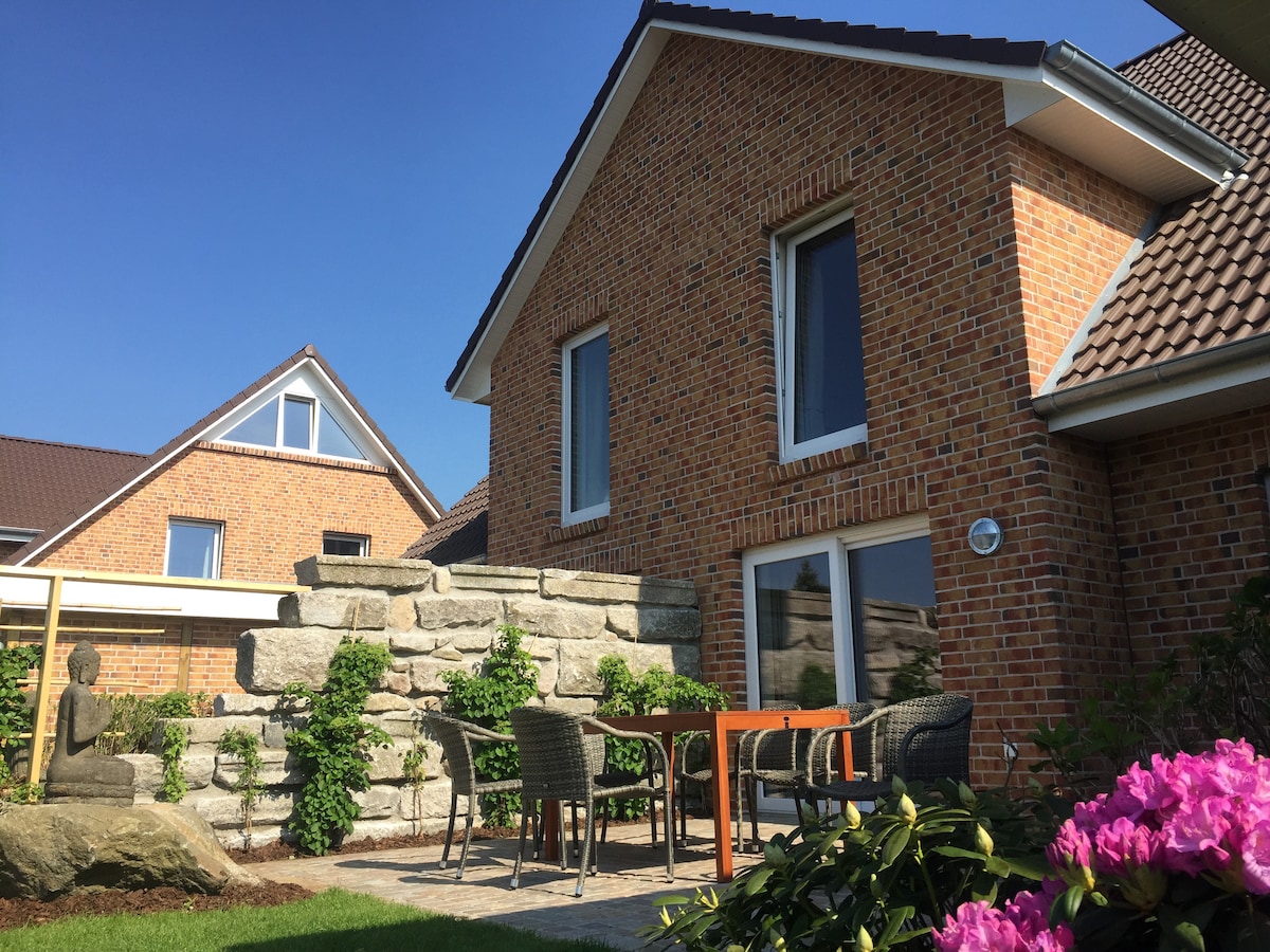 The exterior showcases a modern brick structure with large windows and a unique stone wall. A seating area with wicker chairs and a wooden table is positioned on a stone patio, surrounded by greenery and flowering plants under a clear blue sky.