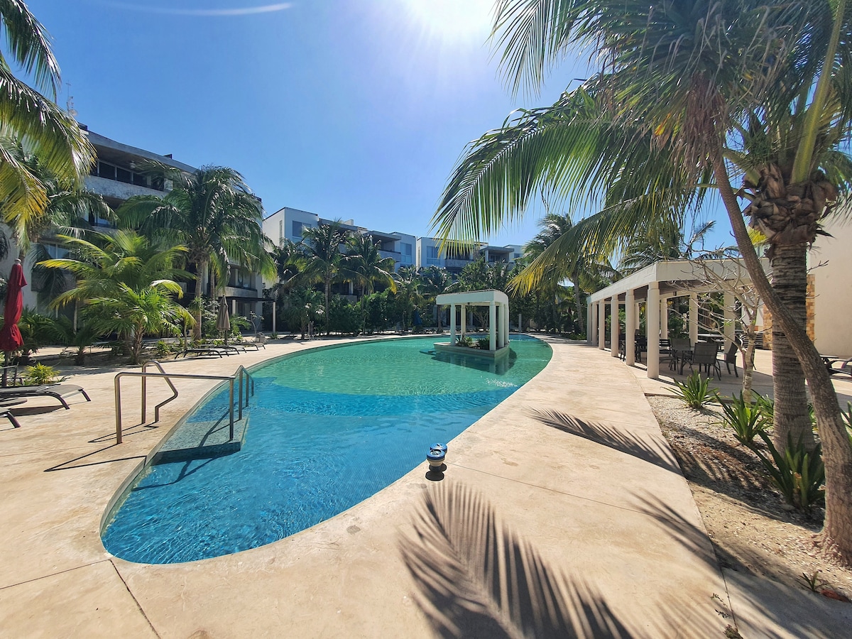 A serene swimming pool is surrounded by lush palm trees and local landscaping. Lounge chairs line one side, offering shaded areas alongside a gazebo situated in the center. The inviting, clear water reflects the blue sky, creating a tranquil atmosphere.