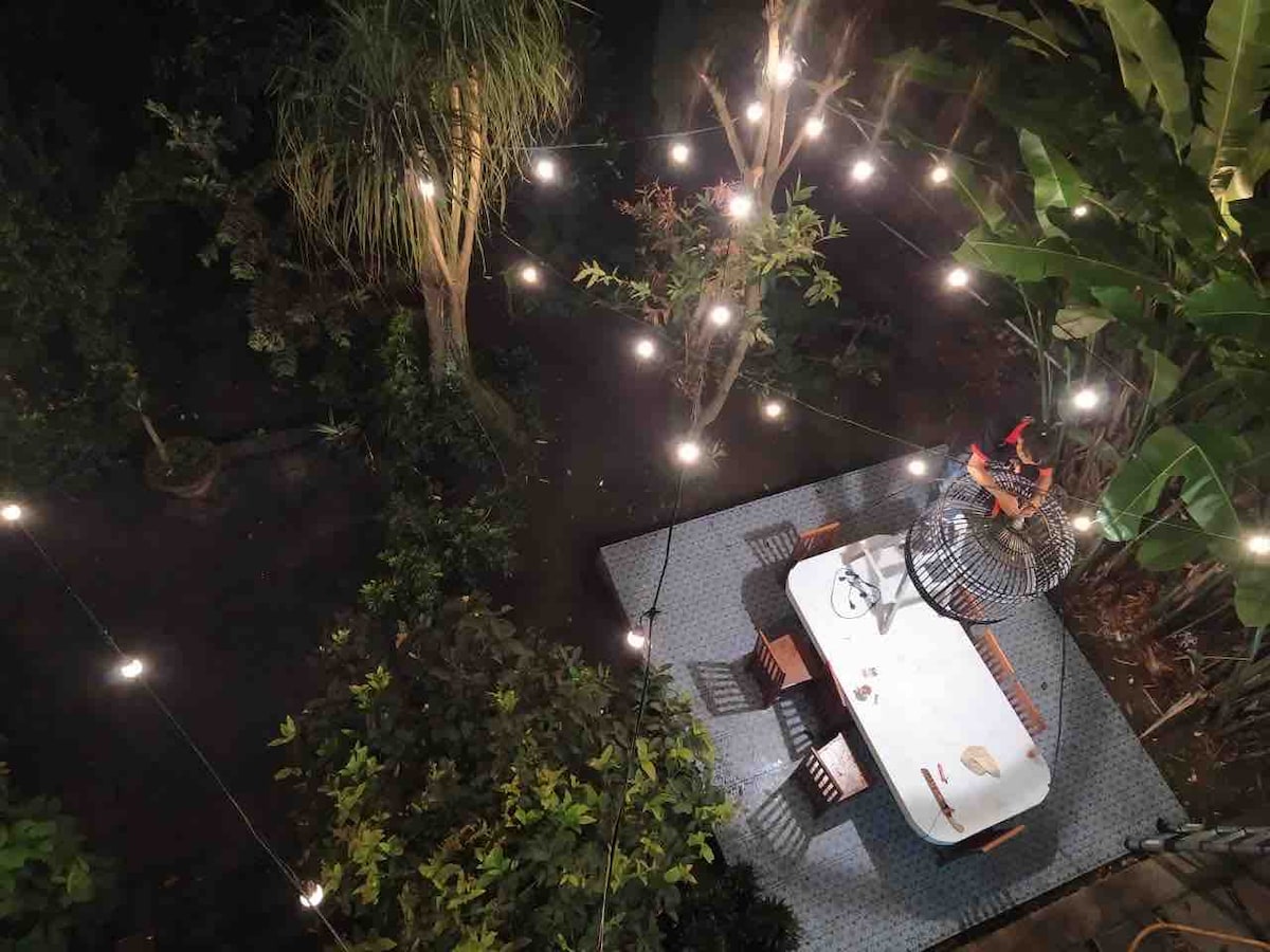 An outdoor dining area is illuminated under a canopy of string lights, surrounded by lush greenery. A large table with several chairs is positioned on a patterned rug, creating an inviting space for gatherings in a serene garden setting.