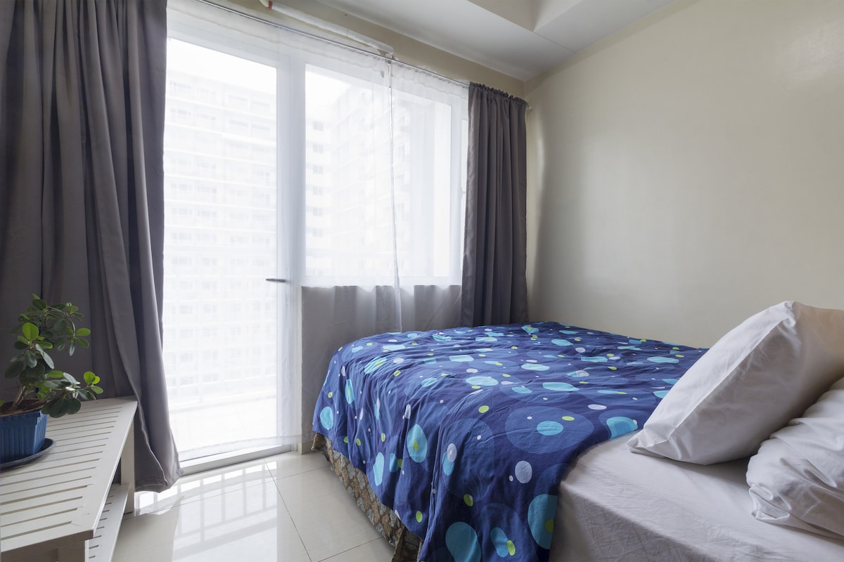 A light and airy bedroom features a queen size bed adorned with a blue polka dot bedspread. Natural light streams through large glass doors, which lead to a balcony. A small potted plant sits on a white table near the bed, enhancing the serene atmosphere.