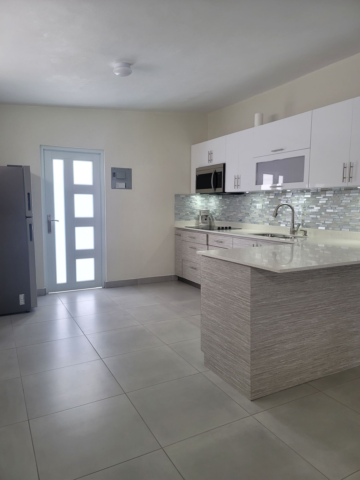 A modern kitchen with sleek cabinetry and a spacious island is depicted. Light-colored granite countertops complement the neutral-toned tile flooring. Natural light fills the area through a glass door, while stainless steel appliances and a stylish backsplash add to the contemporary design.